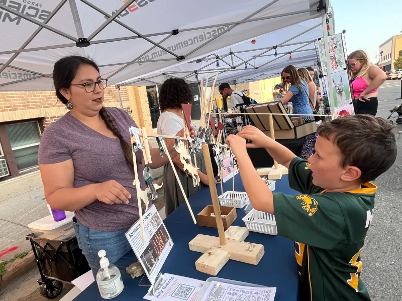 Jessica, an outreach volunteer, is working with a child (in a Packers Jersey) as he places items on the wooden dowel to make both sides balance. It is covered in metal washers and small wood dinosaur cutouts.