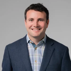 David Schlossman headshot. He has dark brown short wavy hair and is wearing a charcoal blue blazer with a blue and tan and white plaid collared shirt.