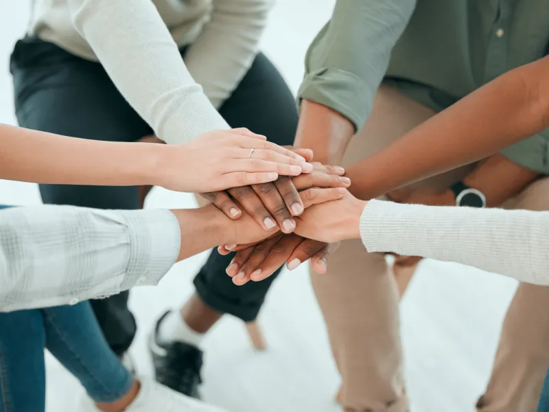 6 adults, of all ages and backgrounds have their hands in the center to signify teamwork.