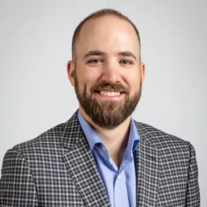 James Hand headshot. He has a mustache and beard and is wearing a blue dress shirt and a grey checked sport coat