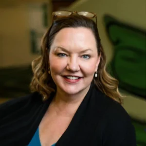 Susan has a dark blazer with a blue shirt. She has long, dark, wavy hair and it is pulled back with sunglasses resting on her head.