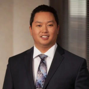 Thad is wearing a dark suit, white shirt, with a multi color tie. He has short, dark hair and is standing in a professional office setting.